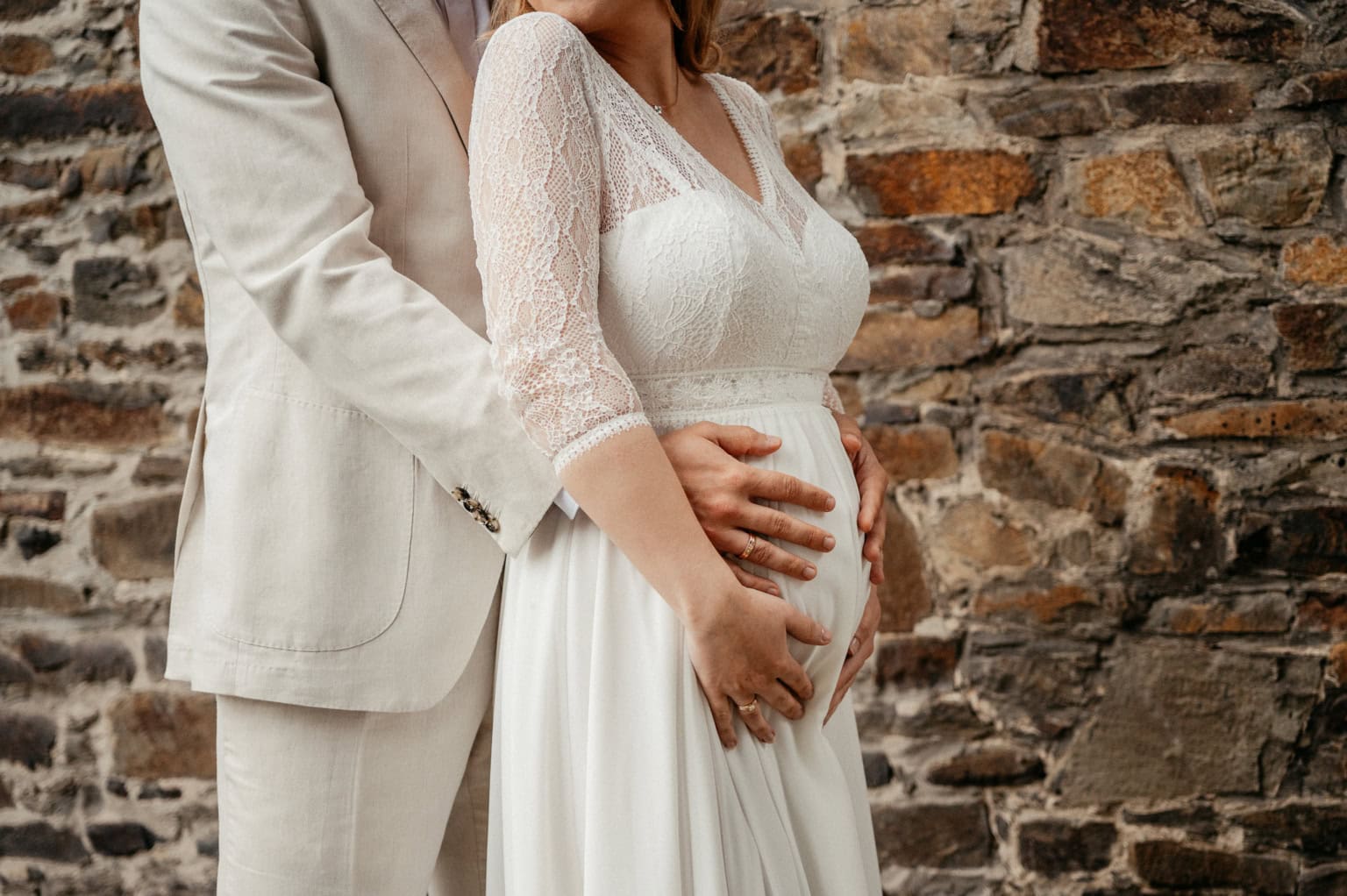 Schwangere Frau in weißem Kleid mit Partner vor Steinmauer.