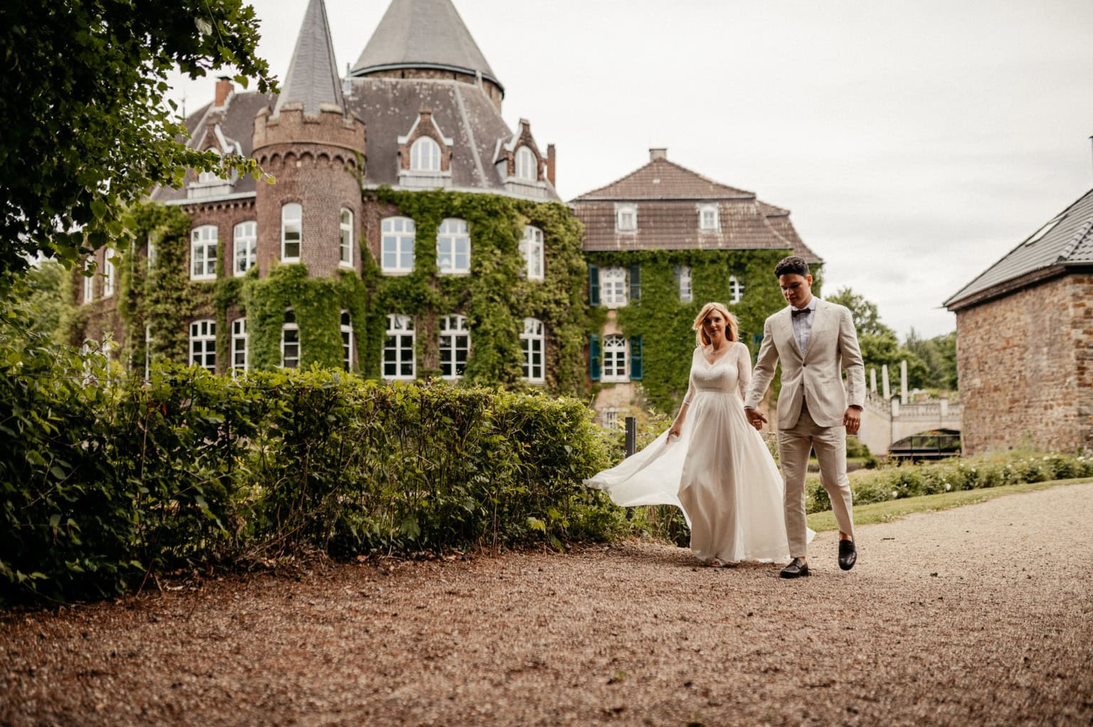 Hochzeitspaar spaziert vor einem historischen Schloss im Grünen.