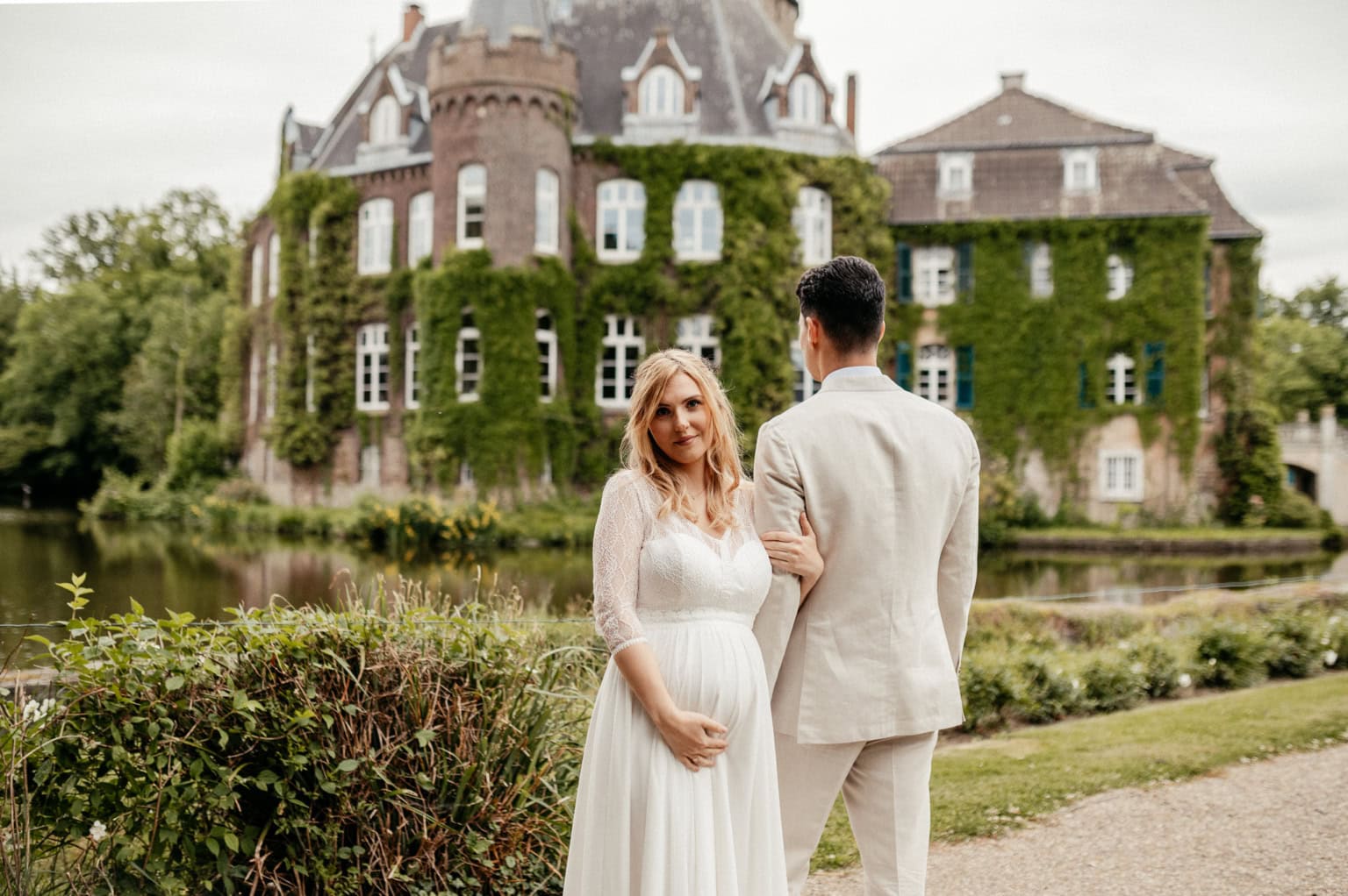 Hochzeitspaar vor einem historischen Schloss, umgeben von Wasser und Natur, bei schönem Wetter.