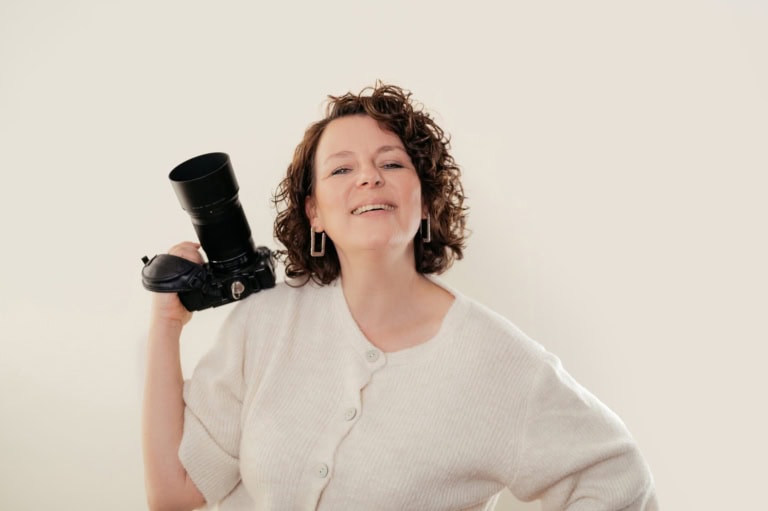 Fotografin mit Kamera, lächelnd und selbstbewusst, vor hellem Hintergrund.