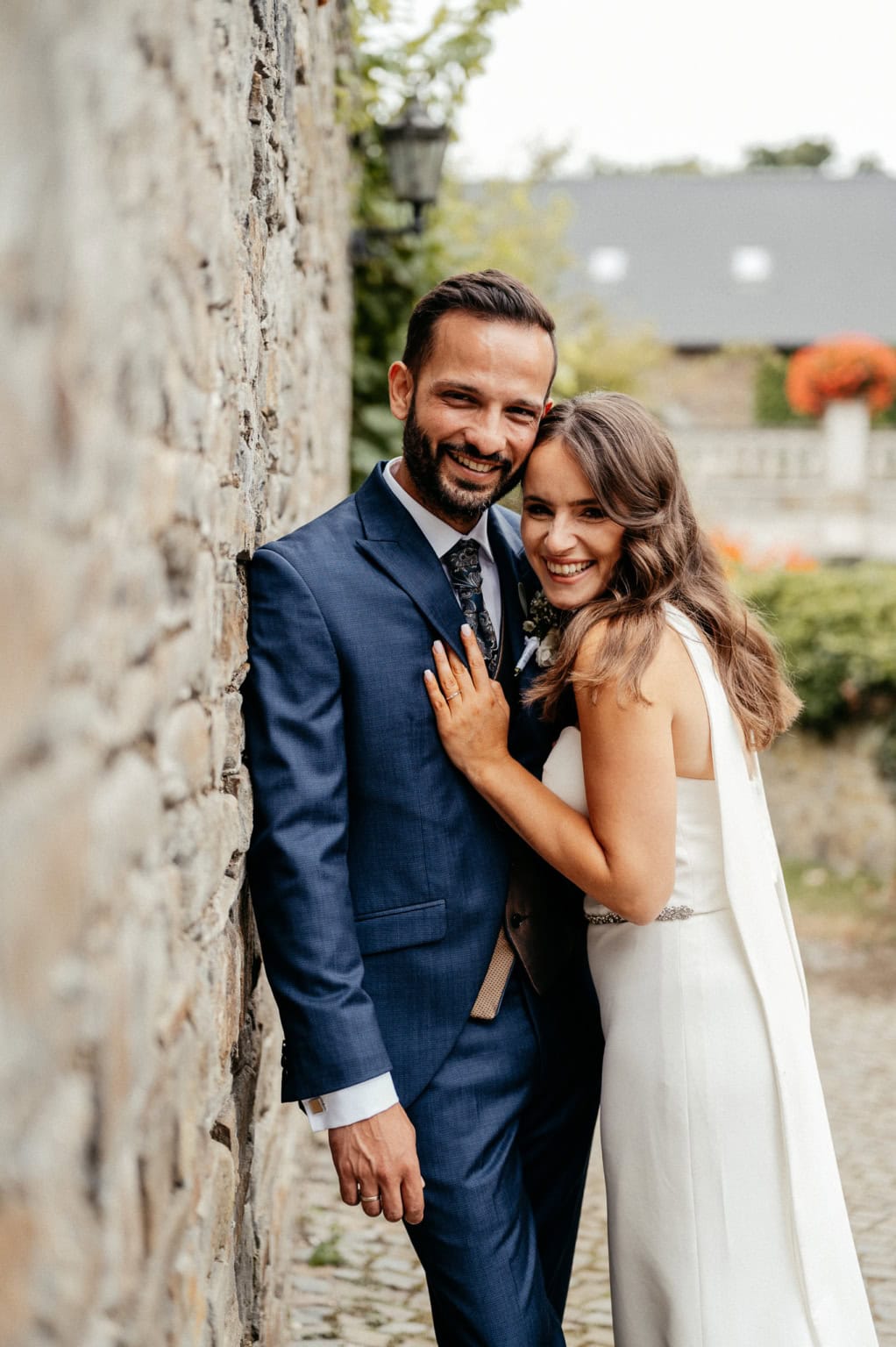 Hochzeitspaar steht eng beieinander, lächelnd und glücklich, im Freien vor einer alten Steinmauer.