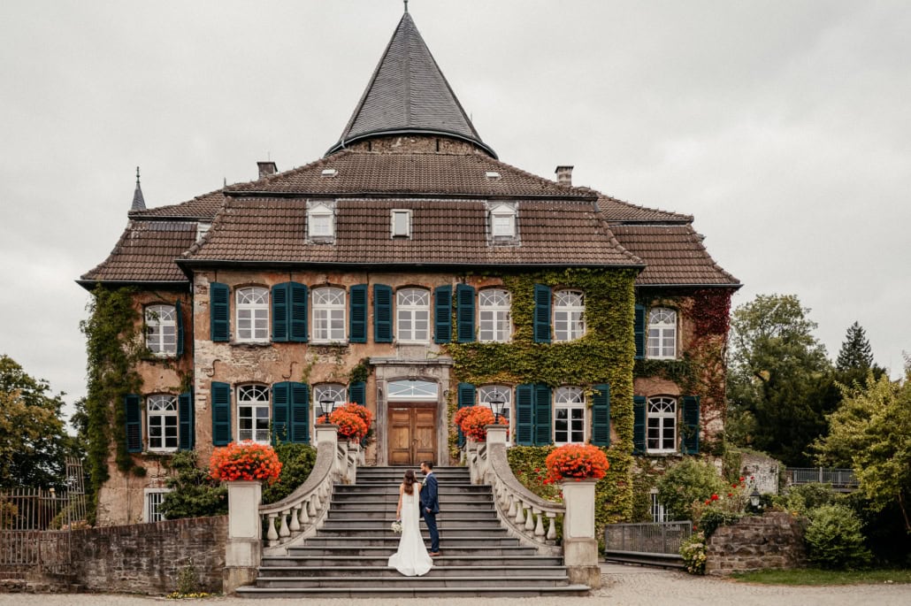 Hochzeitspaar vor einem alten Schloss mit Türmchen, umgeben von bunten Blumen und grünen Bäumen.