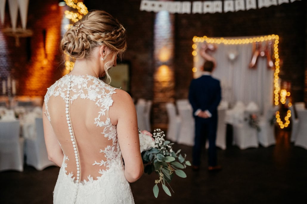 Braut in eleganter Hochzeitskleidung hält Blumenstrauß, Blick auf den Bräutigam im Hintergrund.