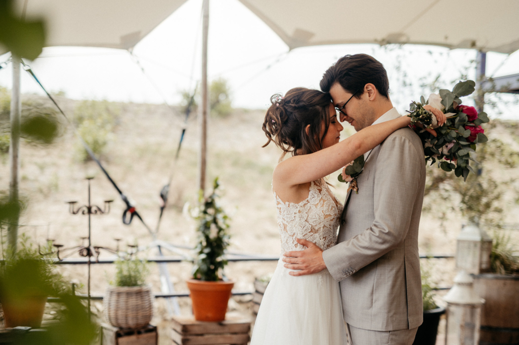 Bride and groom hug closely outdoors, bride in lace dress and bouquet resting over the groom's shoulder.