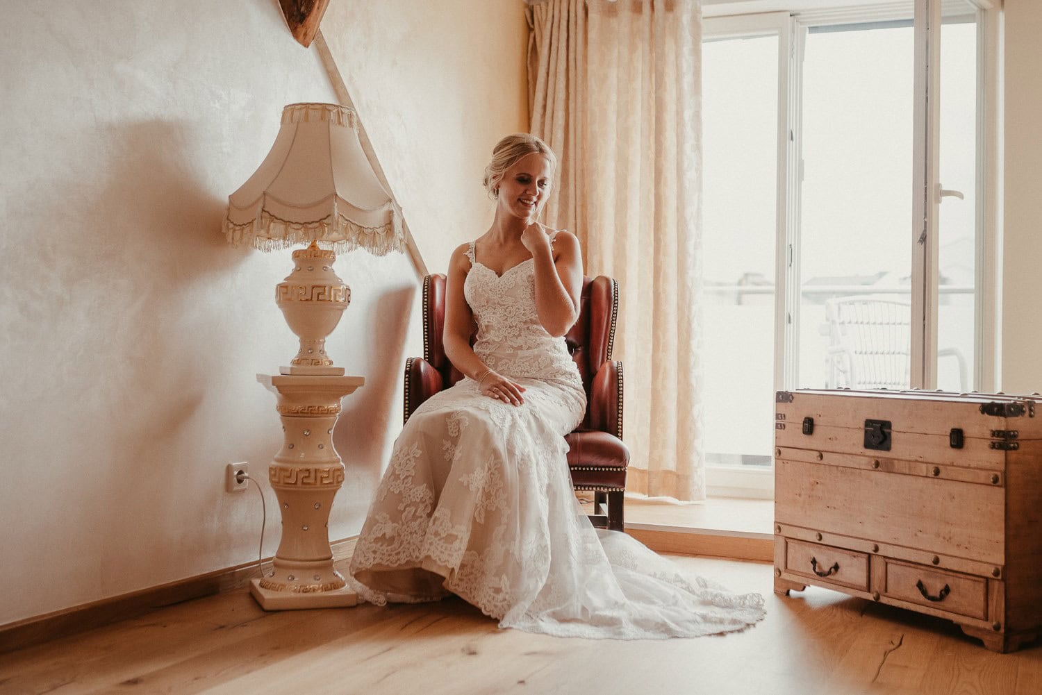 Braut in Hochzeitskleid sitzt entspannt am Fenster, genießt den Moment vor der Zeremonie.