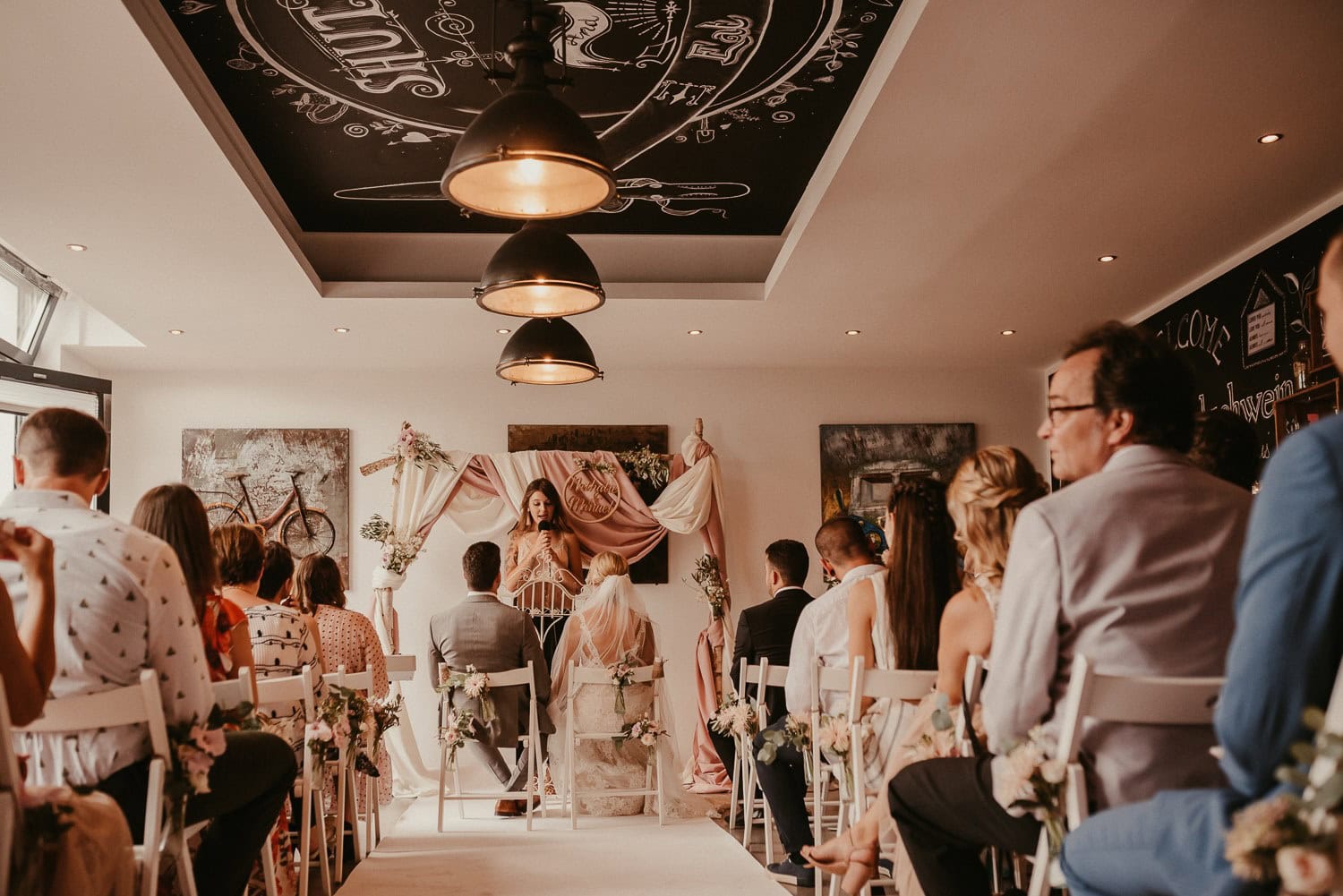 Hochzeitsgäste sitzen im Café während der Zeremonie, mit Blumen und Dekorationen im Hintergrund.