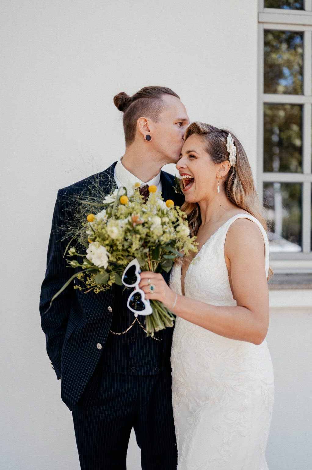 Hochzeitspaar mit Blumenstrauß vor weißem Haus, strahlende Braut und Bräutigam.