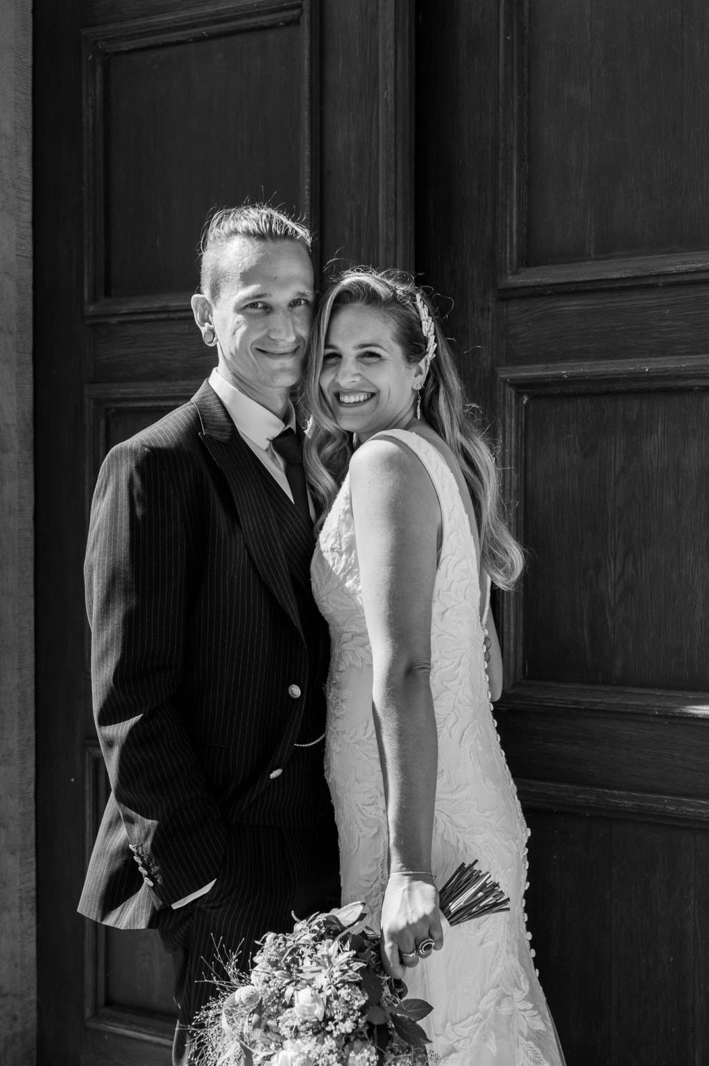 Hochzeitspaar vor dunklen Holztüren, lachend und glücklich, Braut in weißem Kleid, Bräutigam in Anzu.