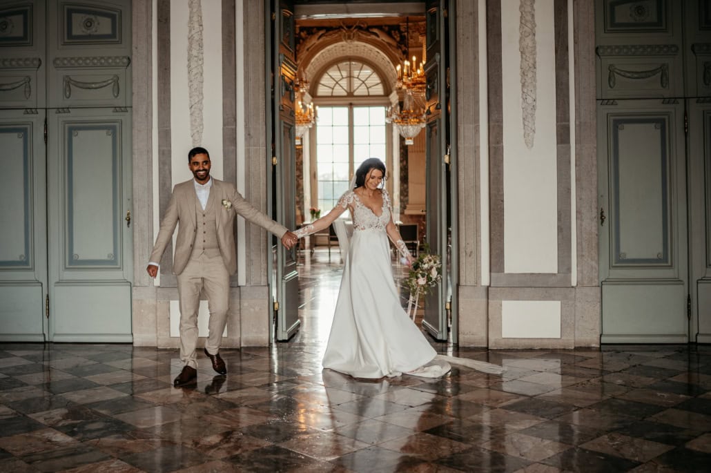 Hochzeitspaar hält Händchen im prunkvollen Saal mit hohen Fenstern und Kronleuchtern.