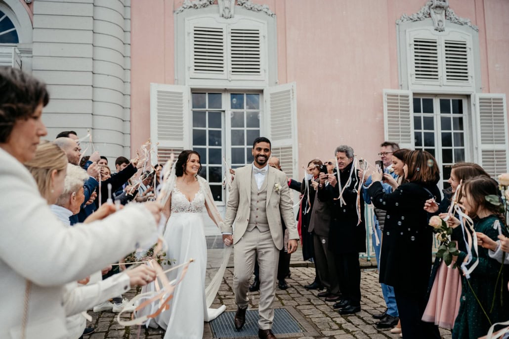 Hochzeitspaar mit Braut in weißem Kleid und Bräutigam in Anzug begrüßen Gäste im Freien.