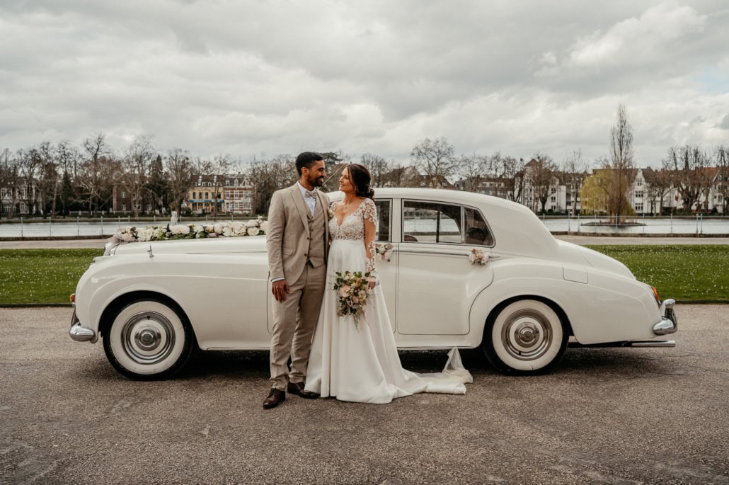 Hochzeitspaar vor einem weißen Oldtimer, das sich liebevoll ansieht, bei bewölktem Himmel.