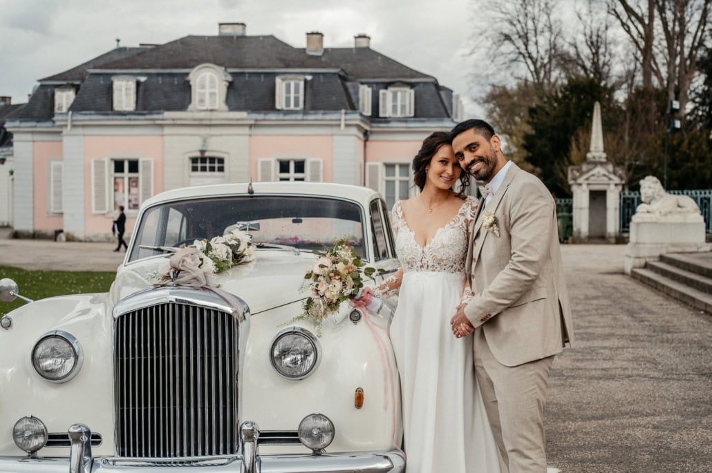 Hochzeitspaar steht neben einem weißen Oldtimer vor einem historischen Gebäude, umgeben von Bäumen u.