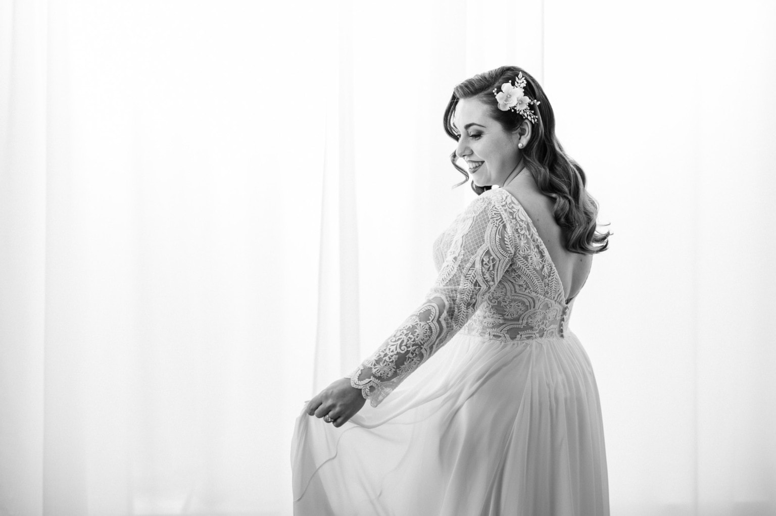 Hochzeitsbraut in einem eleganten weißen Kleid mit Blumen im Haar, lächelnd vor hellem Hintergrund.