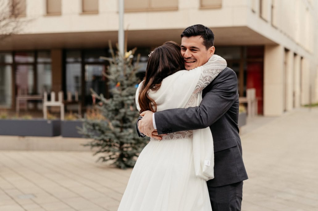 Hochzeitspaar umarmt sich herzlich vor einem modernen Gebäude mit Weihnachtsbaum im Hintergrund.