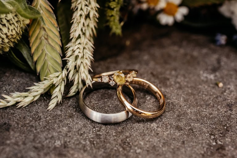 Eheringe und Verlobungsring auf dunklem Steinboden mit Blumen im Hintergrund.