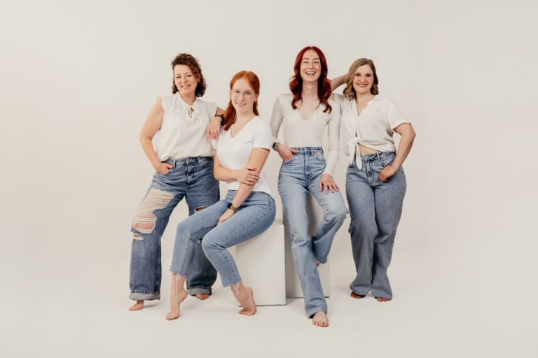Vier Frauen in weißen Oberteilen und Jeans vor neutralem Hintergrund.