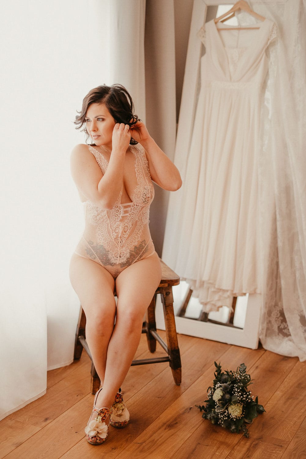 Schöne Frau in zartem Brautkleid sitzt auf einem Stuhl, während sie ihre Haare macht, im Hintergrund.