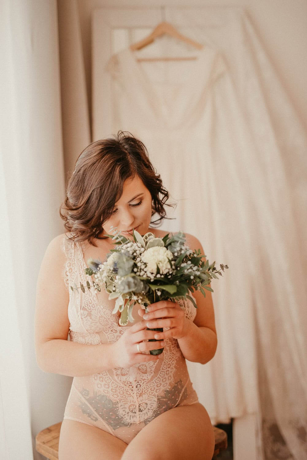 Braut hält Blumenstrauß vor sich, im Hintergrund Brautkleid.