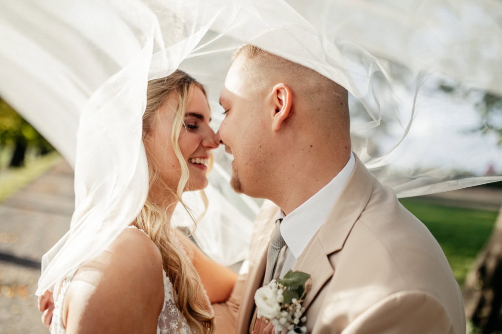 Hochzeitspaar unter einem weißen Schleier, lächelnd und verliebt im Freien.