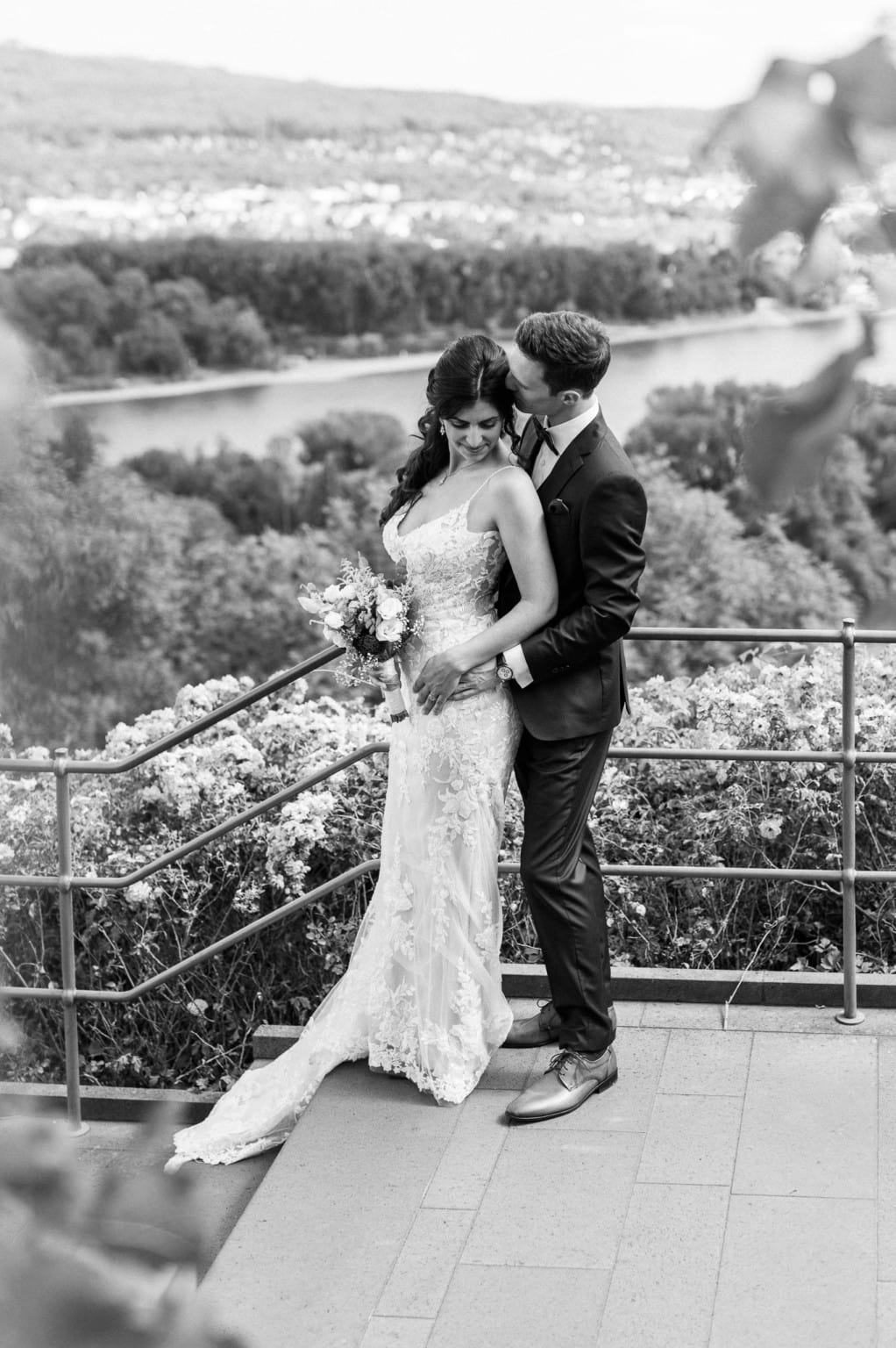 Hochzeitspaar auf Balkon mit Fluss und Natur im Hintergrund, romantisch und elegant.