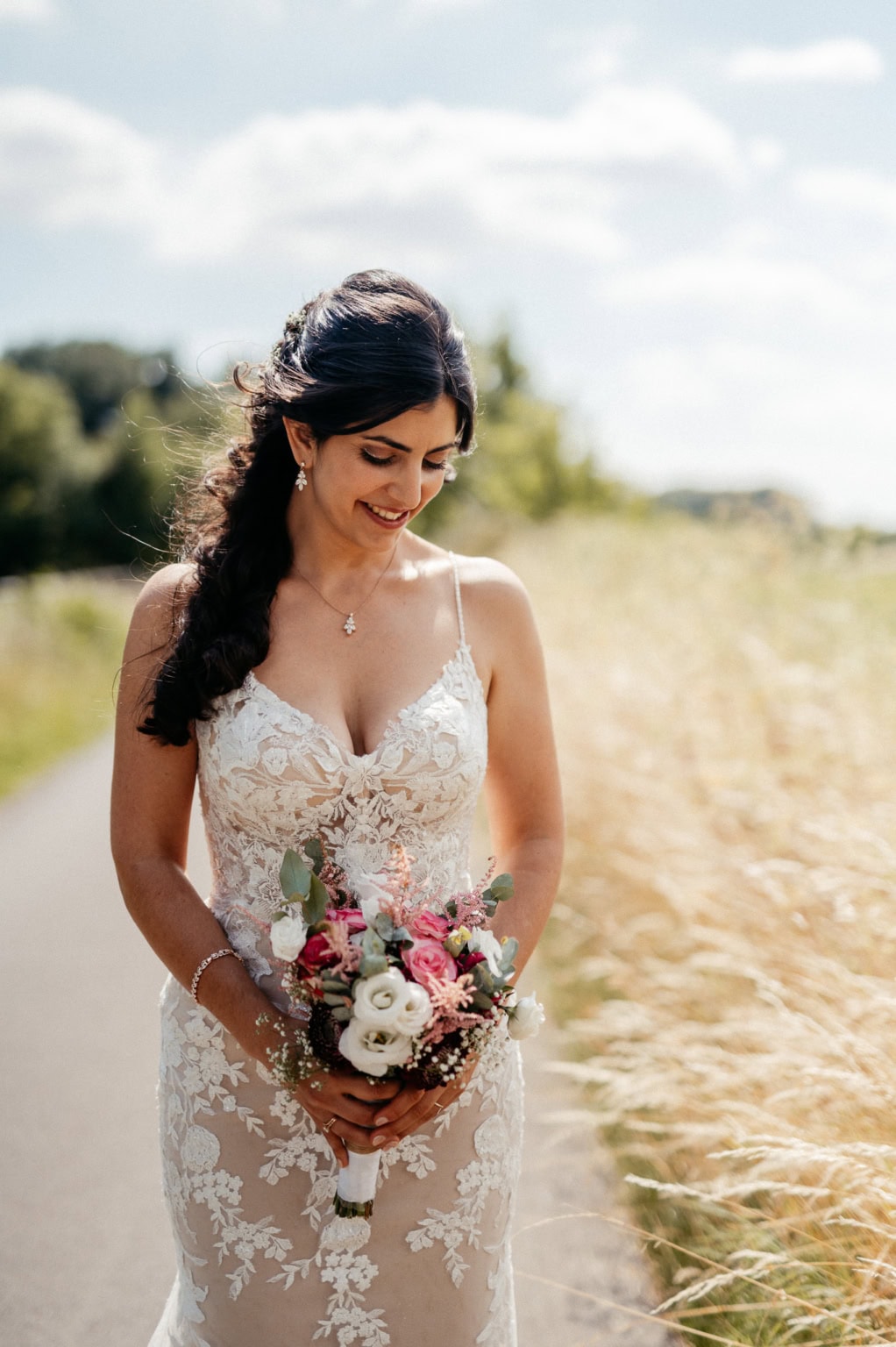 Hochzeitsbraut in einem eleganten Spitzenkleid hält einen Blumenstrauß im Freien bei sonnigem Wett.