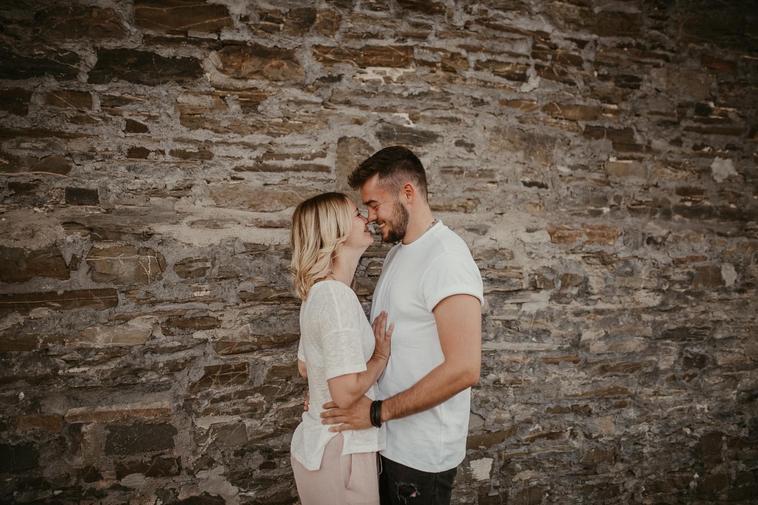 Hochzeitspaar umarmt sich liebevoll vor einer alten Steinmauer.