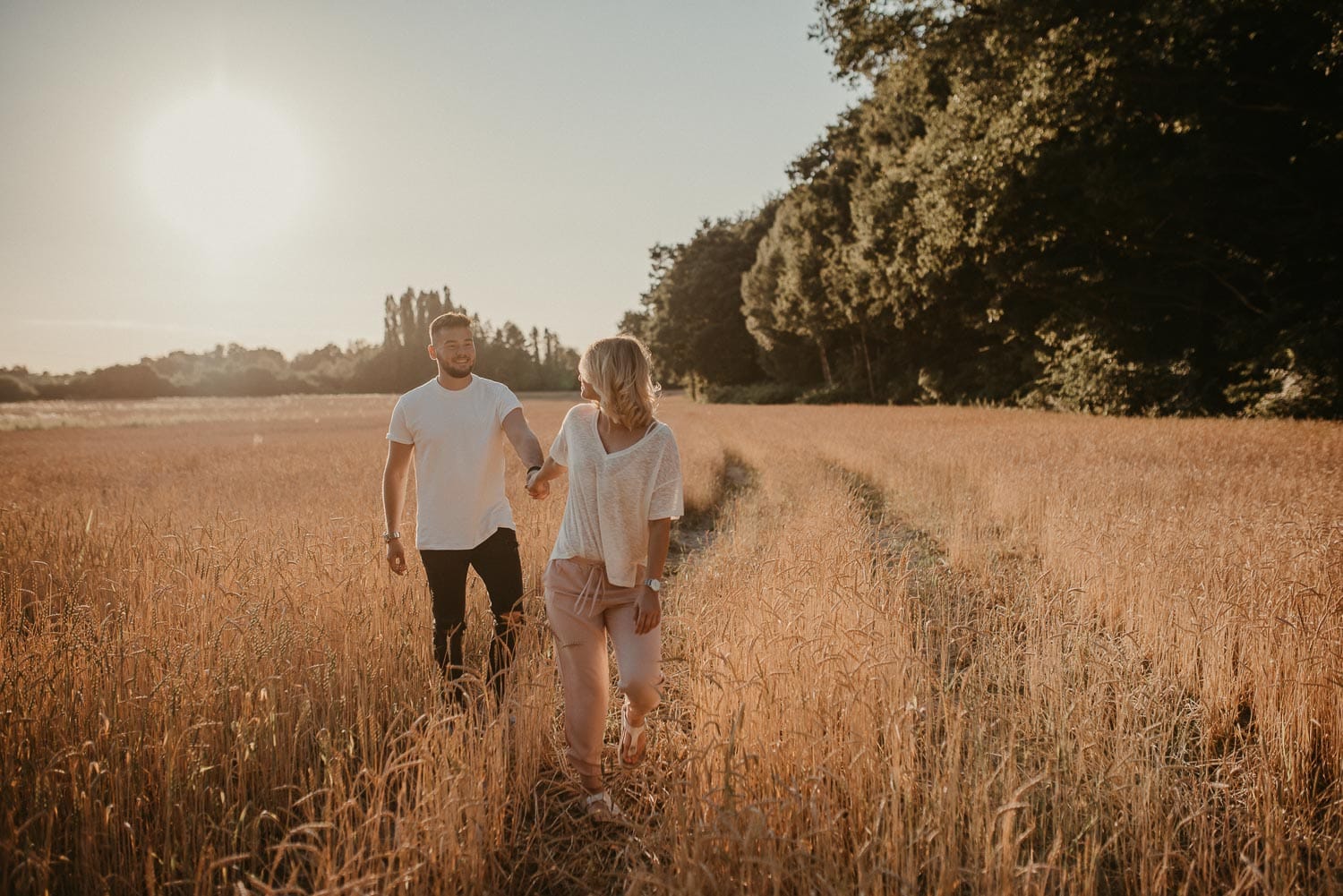 Paar spaziert Hand in Hand durch ein goldenes Getreidefeld bei Sonnenuntergang.
