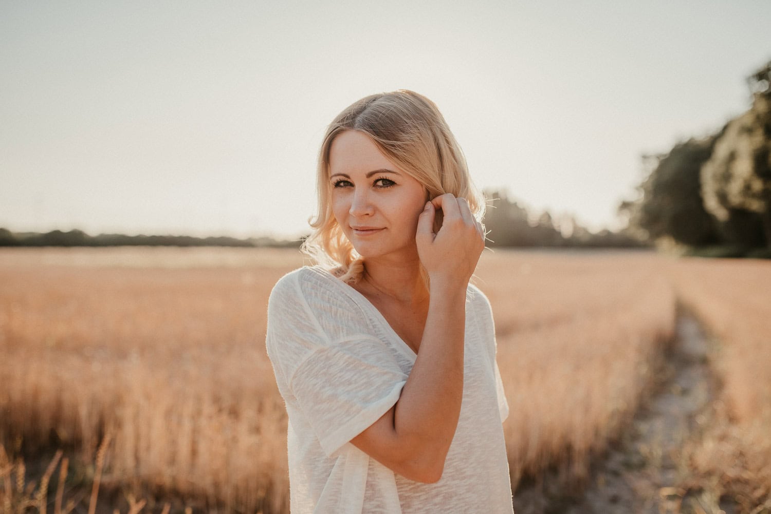 Frau mit blondem Haar in einem weißen Kleid steht in einem goldenen Getreidefeld, Sonnenlicht im Hin.