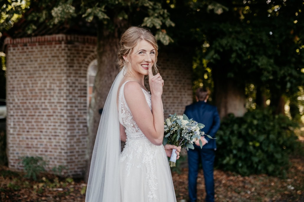 Hochzeit im Garten: Braut mit Blumenstrauß, lächelnd, im Vordergrund, Bräutigam im Hintergrund.